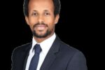 Headshot of Mek Mesfin, who is dressed in a dark suit with a white shirt and a patterned tie. He has short hair and a neatly trimmed beard, and he is looking directly at the camera with a composed expression. The background is plain black.