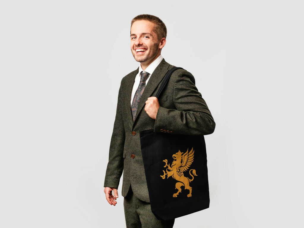 Model wearing a suit with black tote bag with gold logo, on white background