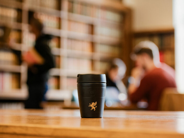 A black reusable coffee cup with a gold emblem of a griffin is placed on a wooden table. The background is blurred, showing people in Gray's Inn library, with bookshelves and individuals working or reading. The focus is on the cup, highlighting its design and placement