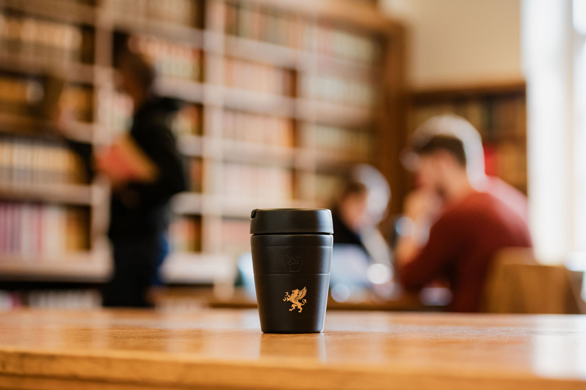 A black reusable coffee cup with a gold emblem of a griffin is placed on a wooden table. The background is blurred, showing people in Gray's Inn library, with bookshelves and individuals working or reading. The focus is on the cup, highlighting its design and placement