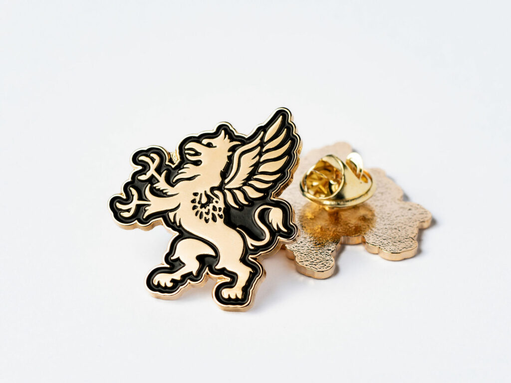 A gold and black enamel pin featuring a detailed griffin is displayed against a plain white background. The griffin, with its wings spread and tail curved, designed with fine lines and shading. The back of the pin is also visible, showing the clasp mechanism and the textured surface