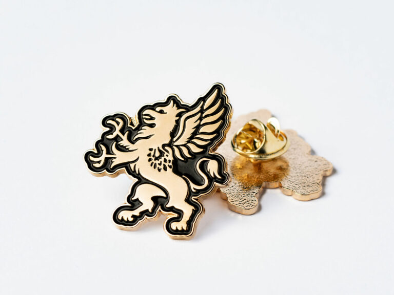 A gold and black enamel pin featuring a detailed griffin is displayed against a plain white background. The griffin, with its wings spread and tail curved, designed with fine lines and shading. The back of the pin is also visible, showing the clasp mechanism and the textured surface