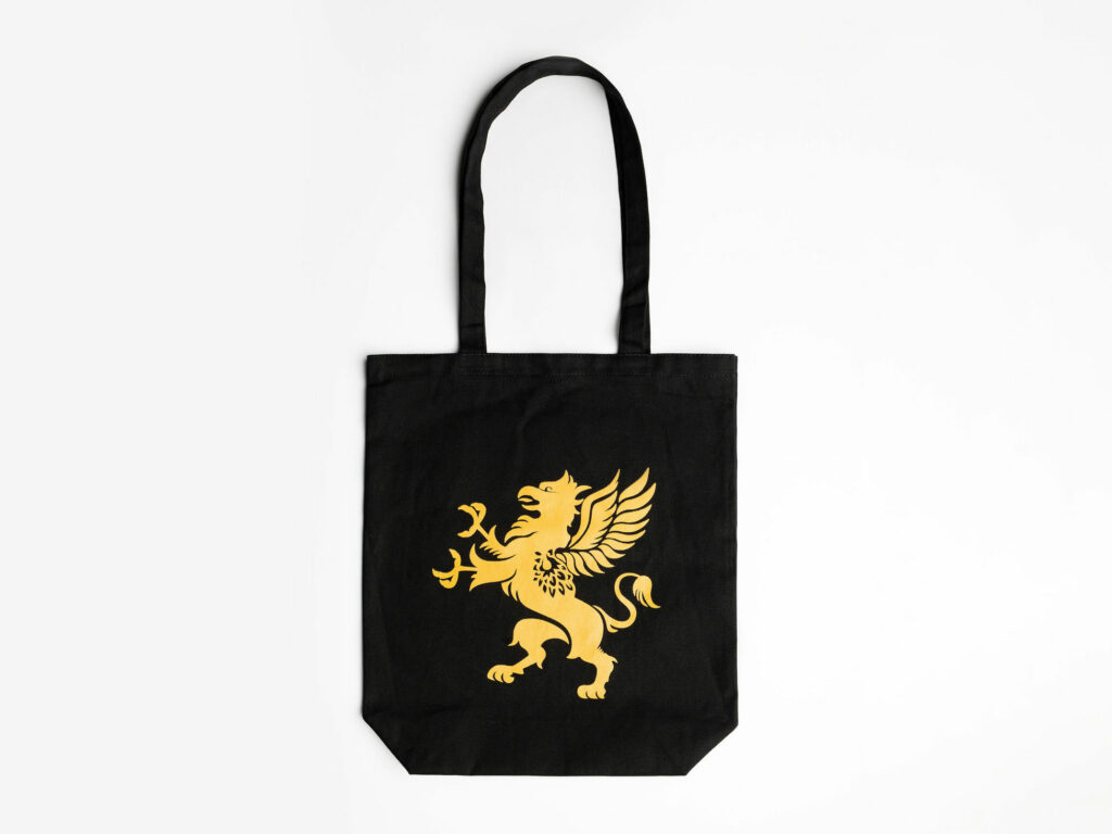 A black tote bag with a gold emblem of a griffin is displayed against a plain white background. The bag features long handles and a simple, elegant design. The griffin emblem stands out prominently