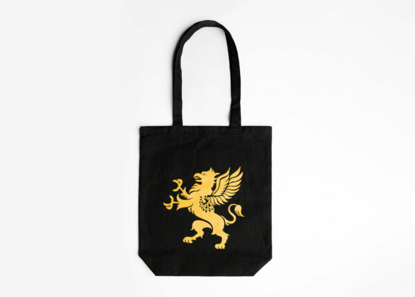A black tote bag with a gold emblem of a griffin is displayed against a plain white background. The bag features long handles and a simple, elegant design. The griffin emblem stands out prominently