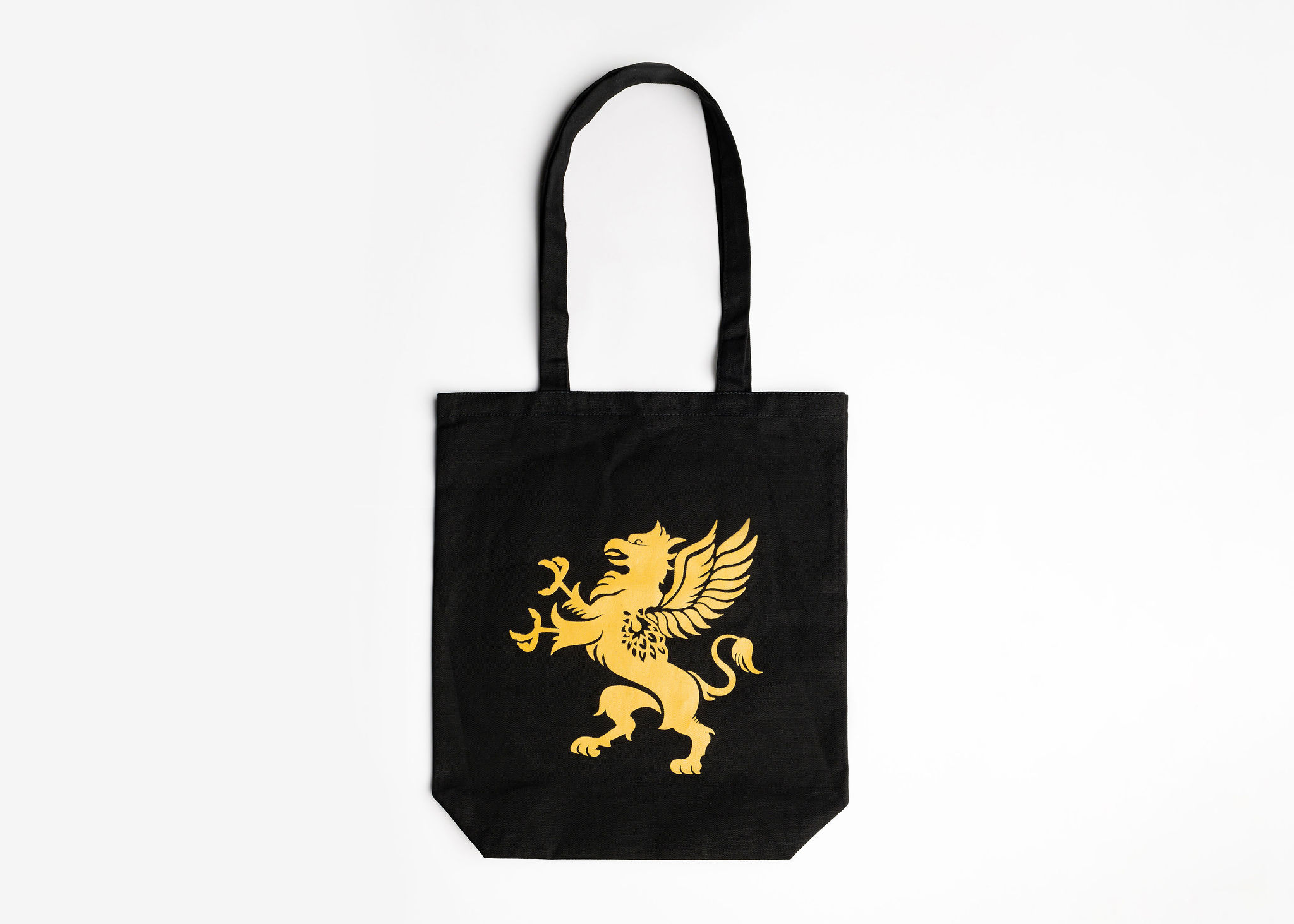 A black tote bag with a gold emblem of a griffin is displayed against a plain white background. The bag features long handles and a simple, elegant design. The griffin emblem stands out prominently
