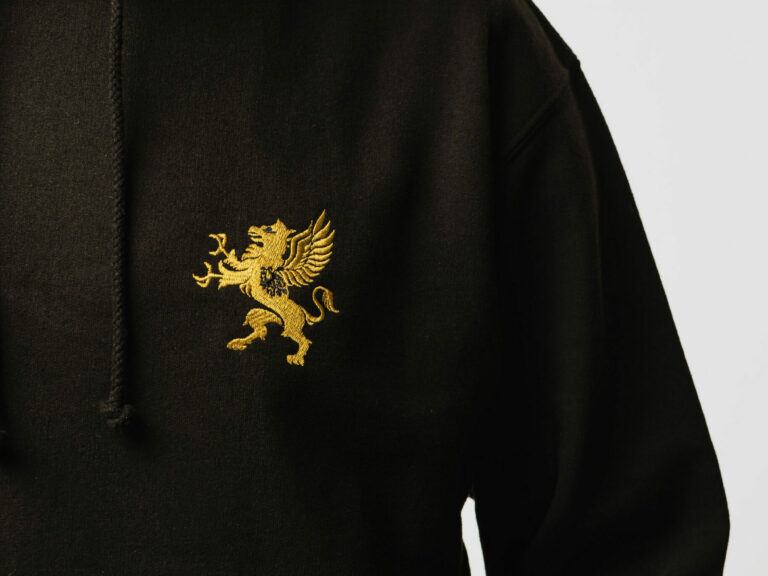A person is wearing a black hoodie with a gold emblem of a griffin embroidered on the chest. The hoodie features a drawstring hood, and the individual's face is not visible in the image