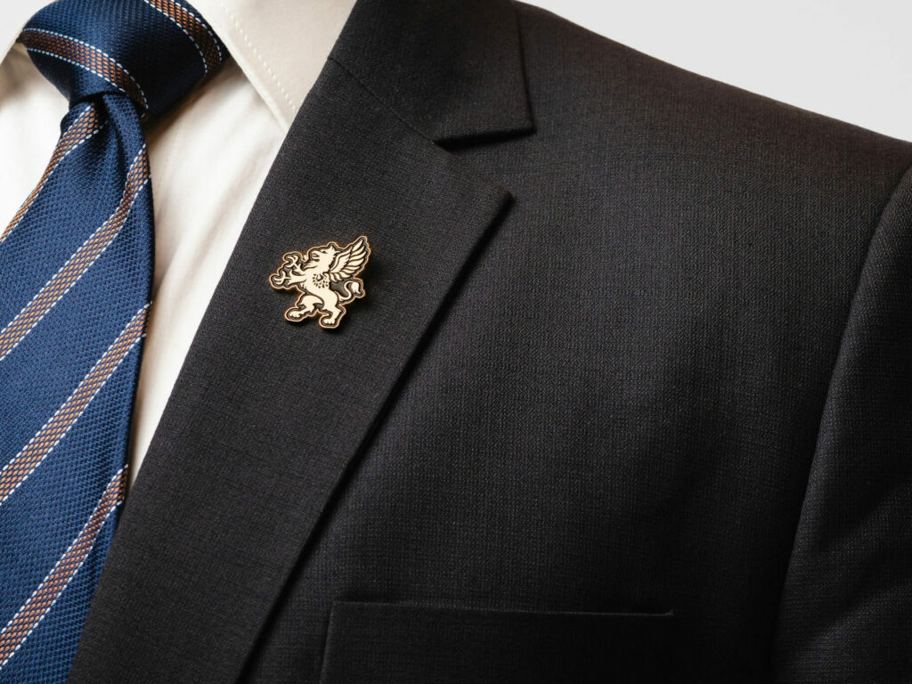A close-up of a man wearing a dark suit with a white shirt and a blue striped tie. The suit jacket features a gold pin with an emblem of a griffin on the lapel. The background is plain white.