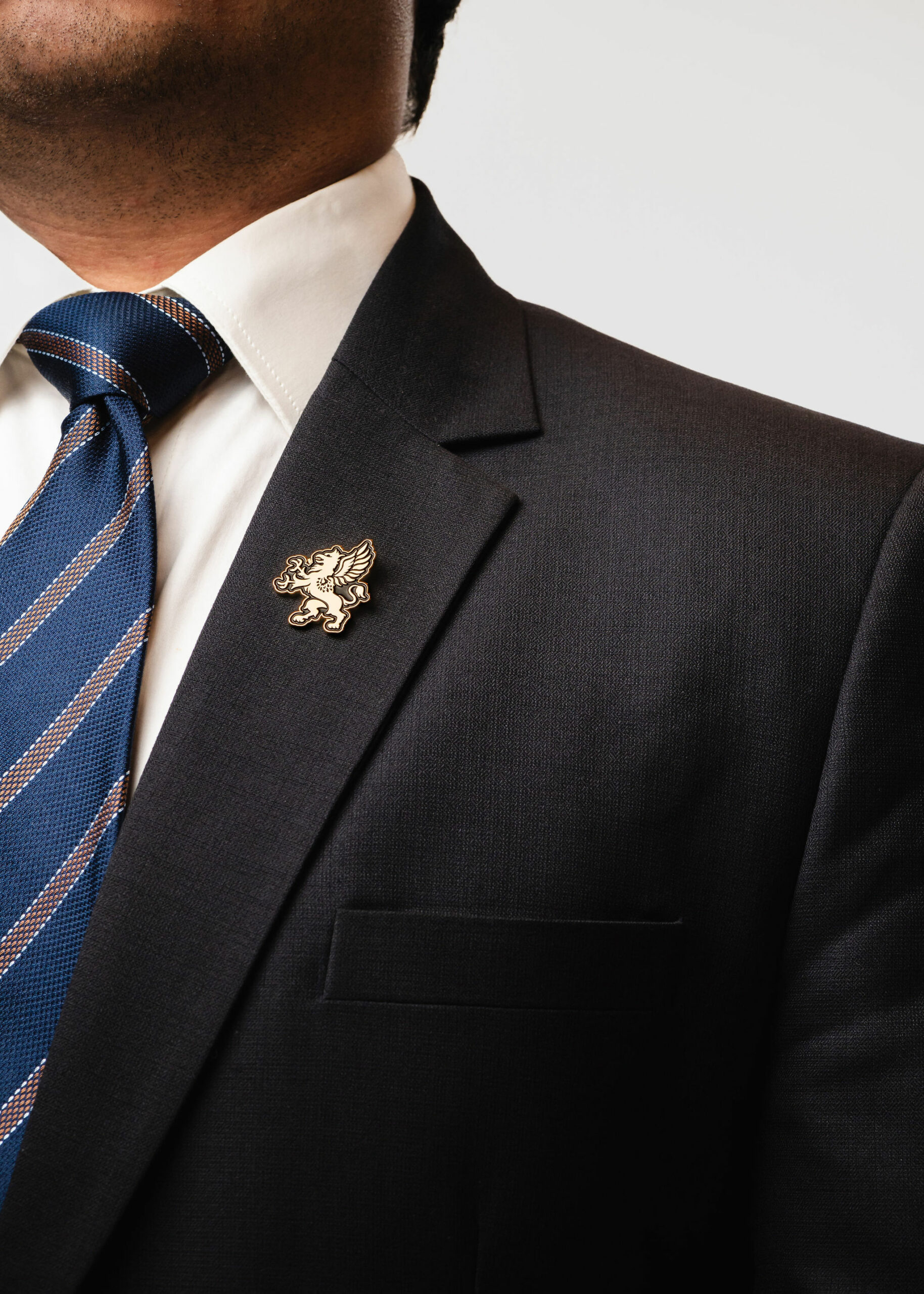 A close-up of a man wearing a dark suit with a white shirt and a blue striped tie. The suit jacket features a gold pin with an emblem of a griffin on the lapel. The background is plain white.