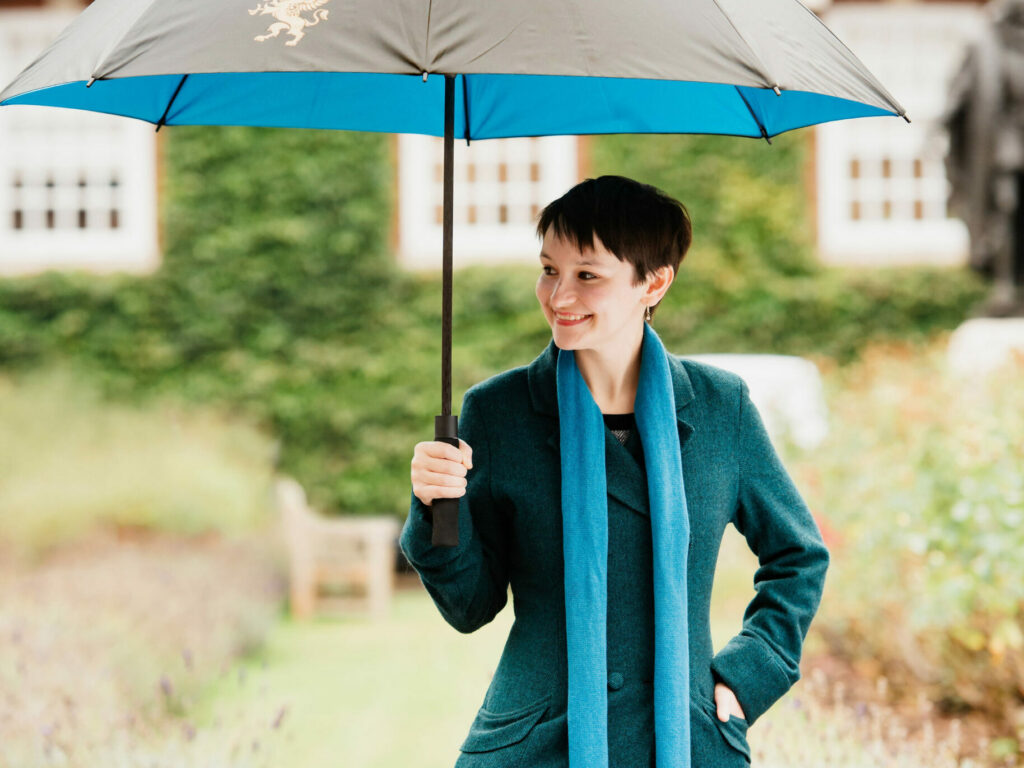 A young woman is standing outdoors, holding a large umbrella with a blue and black design. One panel of the umbrella has a gold griffin on it. She is dressed in a teal coat and matching scarf, smiling warmly. The background features a building with white windows and greenery