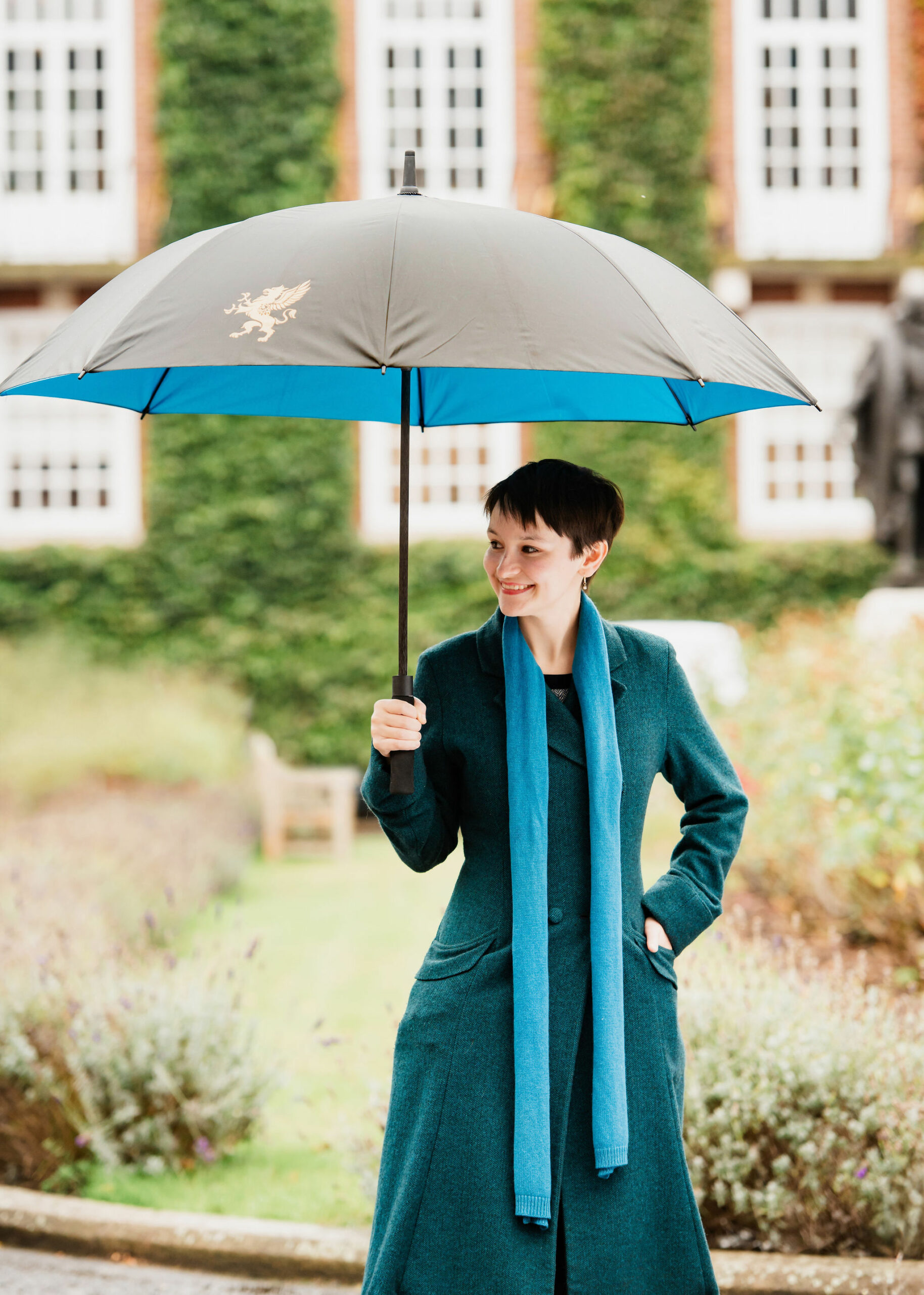 A young woman is standing outdoors, holding a large umbrella with a blue and black design. One panel of the umbrella has a gold griffin on it. She is dressed in a teal coat and matching scarf, smiling warmly. The background features a building with white windows and greenery