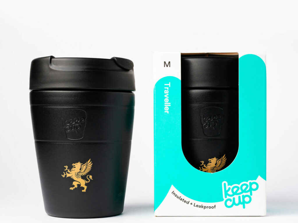A black KeepCup "Traveller" reusable coffee cup is displayed next to its packaging. The cup features a gold griffin emblem on the front. The packaging is predominantly white with teal accents, including the words "Traveller," "Insulated + Leakproof," and "KeepCup." A window in the box shows the same gold griffin logo visible on the cup inside. The size "M" is indicated at the top of the box. Both the cup and the box are set against a clean, white background.