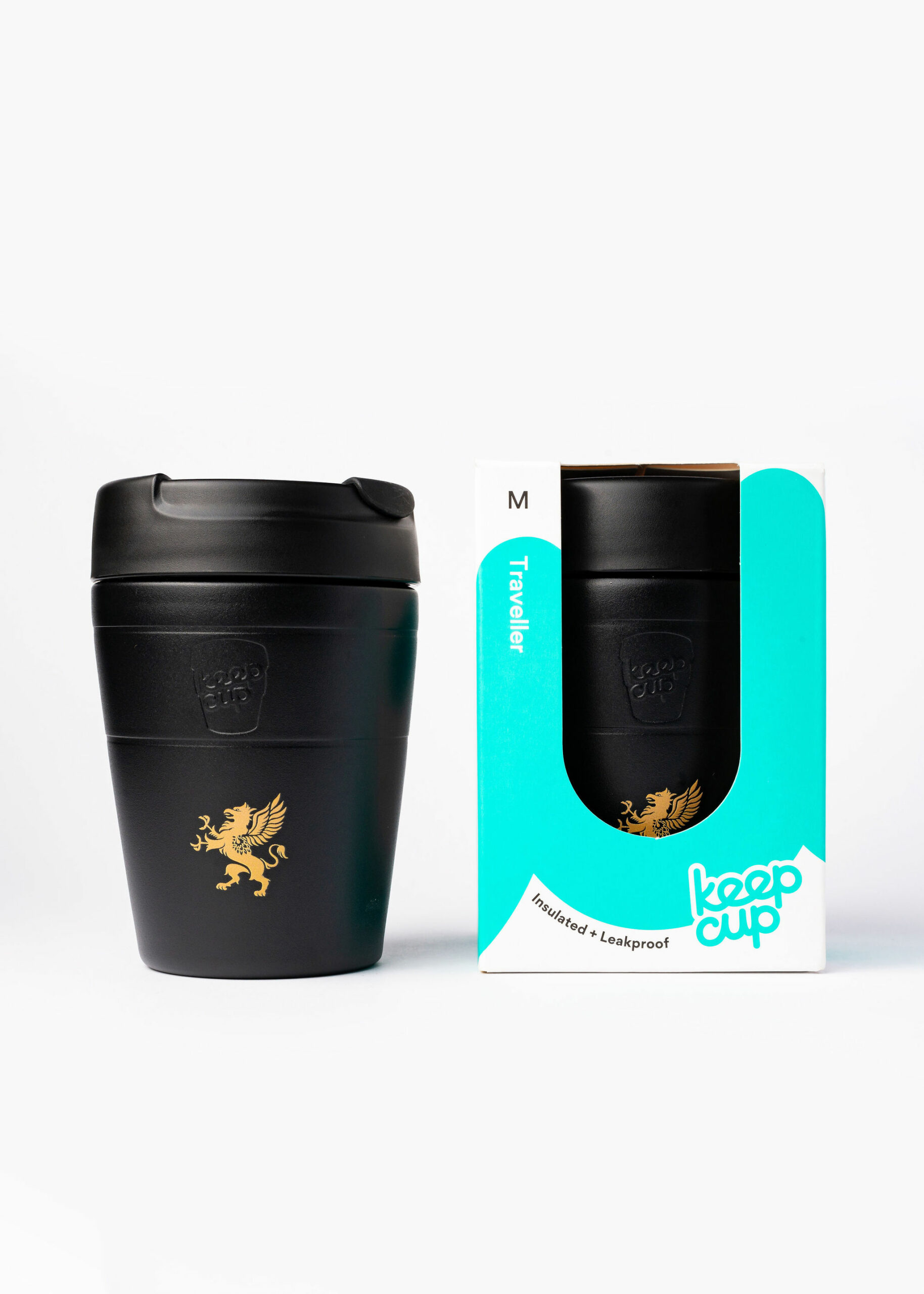 A black KeepCup "Traveller" reusable coffee cup is displayed next to its packaging. The cup features a gold griffin emblem on the front. The packaging is predominantly white with teal accents, including the words "Traveller," "Insulated + Leakproof," and "KeepCup." A window in the box shows the same gold griffin logo visible on the cup inside. The size "M" is indicated at the top of the box. Both the cup and the box are set against a clean, white background.