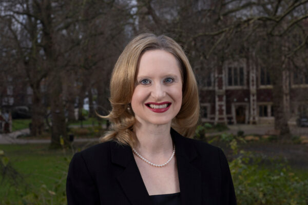 Sara George pictured smiling warmly at the camera. She has shoulder-length blonde hair and is wearing a black blazer with a pearl necklace. The background features an outdoor setting with bare trees and a brick building