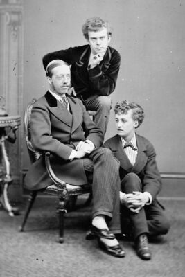 Three people dressed in male Victorian clothing. Two seated and one stood in the middle.