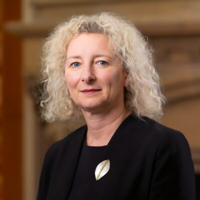 Portrait shot of a woman with blond shoulder length hair wearing a black suit and a brooch