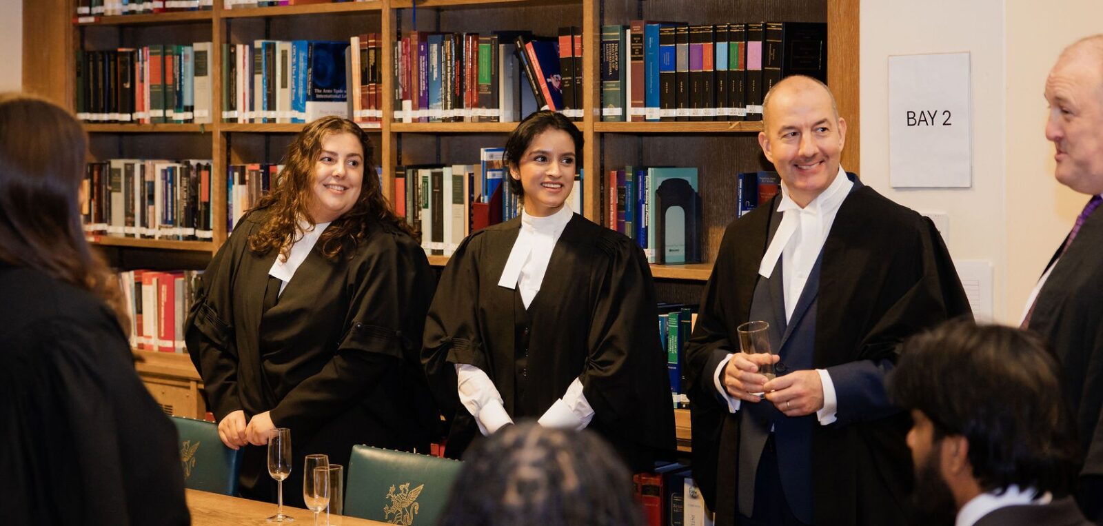A group of callees in barristers robes standing talking and smiling with book shelves in the Library in the background